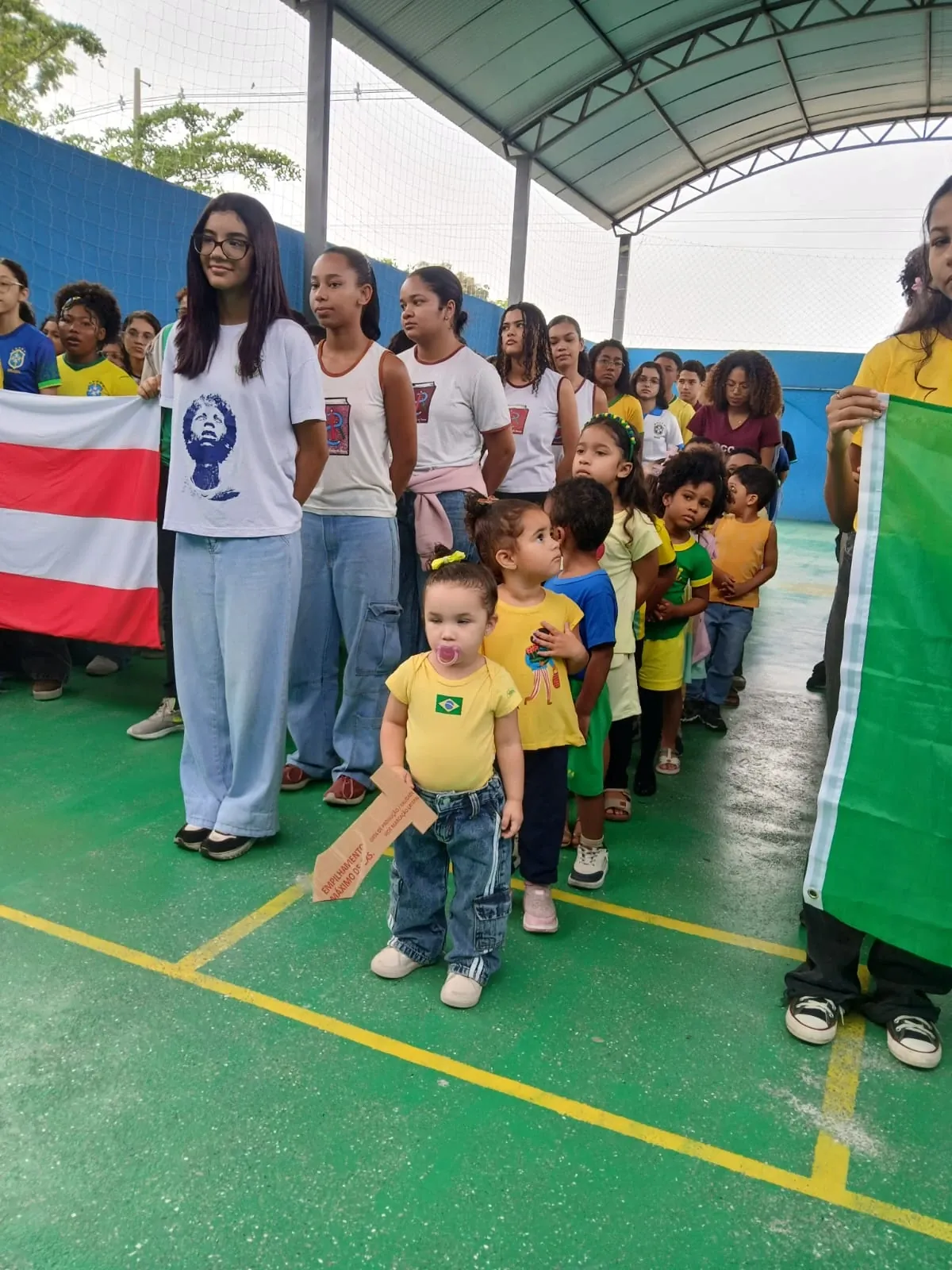Criança pequena em fila com os colegas maiores durante o momento cívico, vestida de verde e amarelo