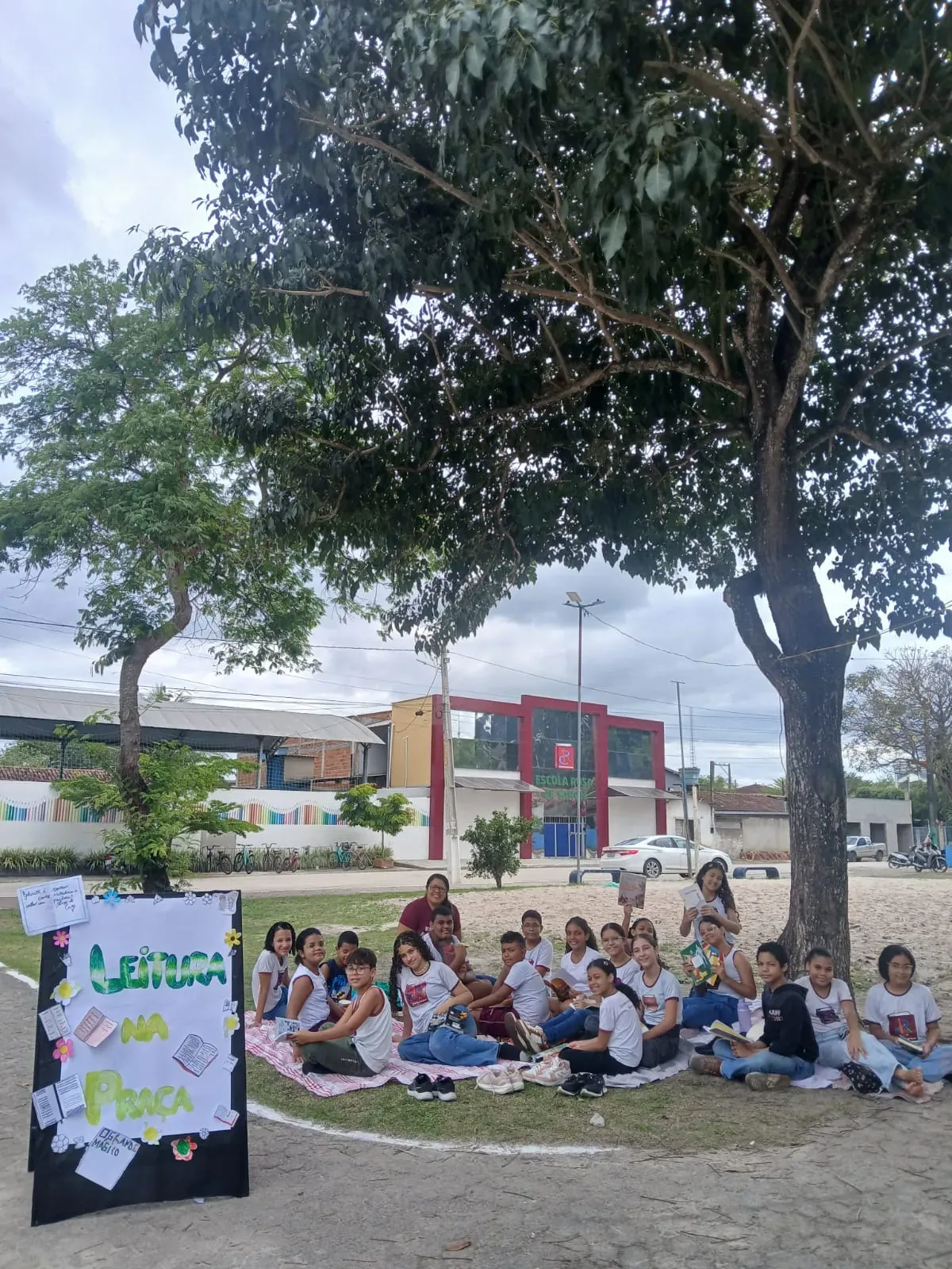 Alunos da Rosa de Saron sentados na praça com livros, em frente à escola, no Leitura na Praça